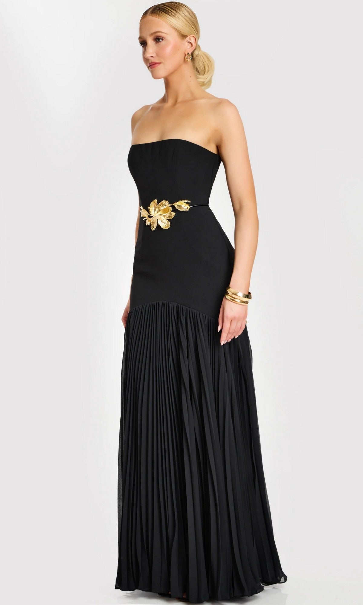 A woman wearing the Jovani 47000 long formal dress—a strapless, floor-length black gown featuring a gold floral waist detail and a pleated skirt—stands against a plain light background.